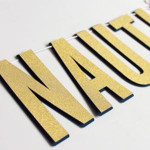 May include: A nautical-themed banner spelling out the word "NAUTI" in gold glitter letters with navy blue edges. Red and white anchor accents are on either side of the banner, strung on a white string.
