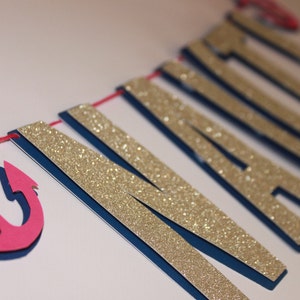 May include: A banner with the word "MARRIED" in gold glitter letters, blue backing, and a pink anchor. The letters are strung on a red string, perfect for a wedding or nautical-themed event.