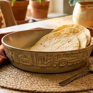 Pode incluir: Uma tigela de cerâmica bege cheia de tortillas, com um padrão decorativo na borda. Uma colher de prata repousa sobre um tapete de tecido. A cena inclui uma pequena estátua e um pano cor de terracota.