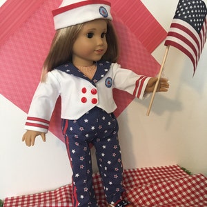 May include: A doll dressed in a sailor outfit with a white hat, jacket, and pants. The outfit has red and blue accents with star patterns. The doll is holding a small American flag.