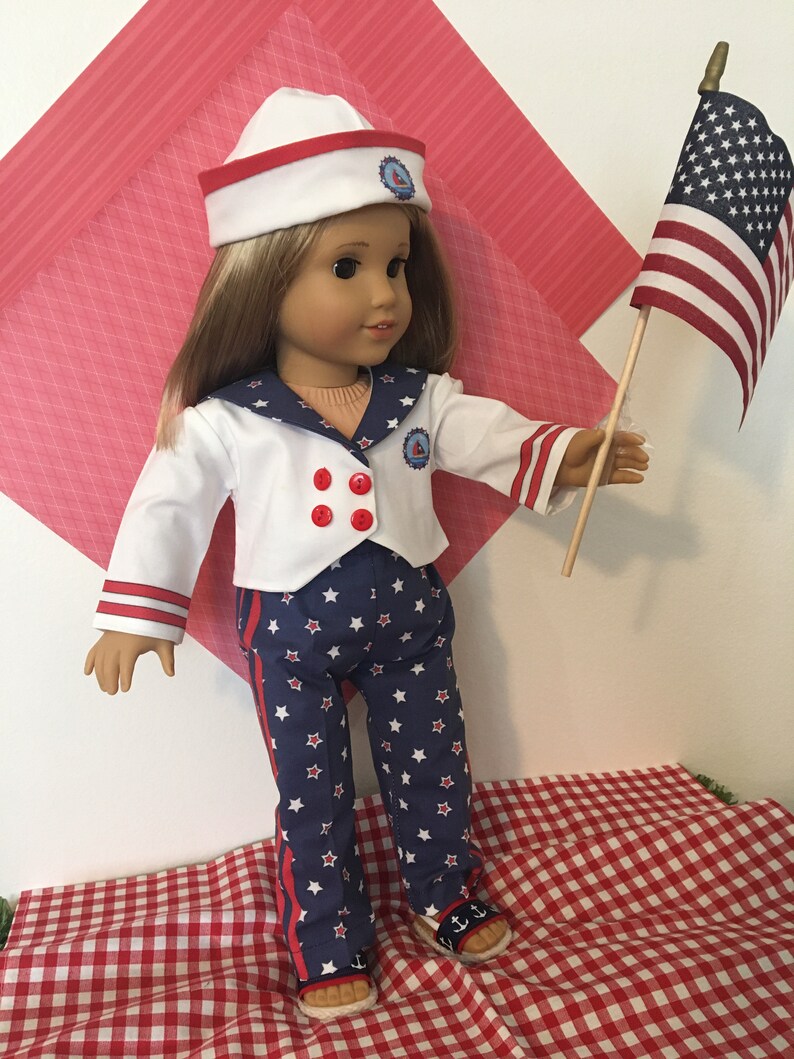 May include: A doll dressed in a sailor outfit with a white hat, jacket, and pants. The outfit has red and blue accents with star patterns. The doll is holding a small American flag.