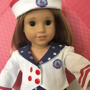 May include: A doll wearing a sailor-themed outfit. The outfit includes a white jacket with red buttons, a navy blue skirt with white stars, and a matching sailor hat. The hat and jacket feature a nautical emblem.
