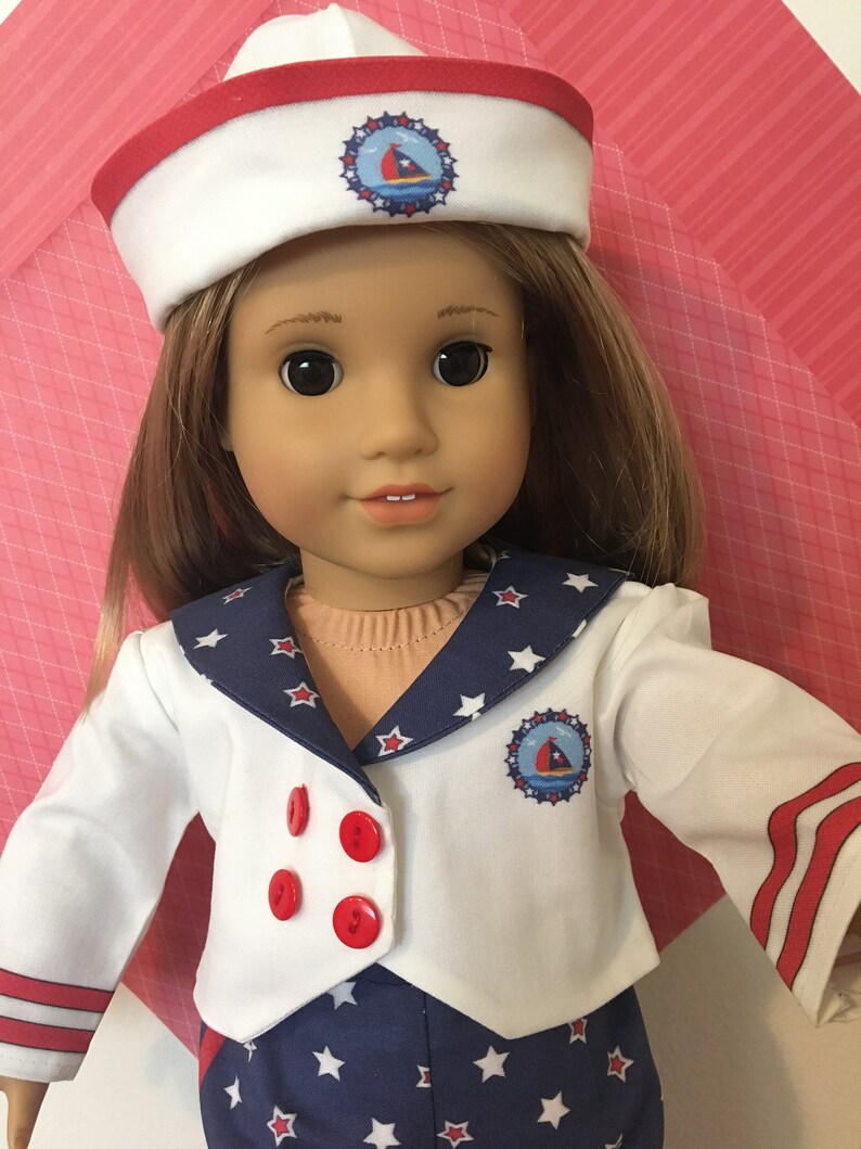 May include: A doll wearing a sailor-themed outfit. The outfit includes a white jacket with red buttons, a navy blue skirt with white stars, and a matching sailor hat. The hat and jacket feature a nautical emblem.