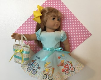 May include: A doll wearing a light blue dress with a bicycle design and a yellow bow. The dress has a light blue ribbon at the waist and a matching purse. The doll is wearing yellow shoes.