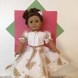 May include: A doll wearing a white dress with gold glitter Christmas tree and snowman designs. The dress has short, puffy sleeves with gold trim and a gold ribbon belt. The doll has brown hair and blue eyes, and is wearing beige shoes.