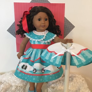 May include: A doll wearing a turquoise dress with a penguin and snowflake pattern, a red ribbon, and matching shoes. A matching cape is displayed on a stand. The doll has dark curly hair and is posed in front of a red and gray backdrop.