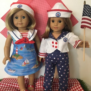 May include: Two dolls dressed in nautical-themed outfits. One doll wears a blue dress with a sailboat print, a red bow, and a white sailor hat. The other doll is in a white jacket, blue star-patterned pants, and a sailor hat. An American flag is held by one doll.
