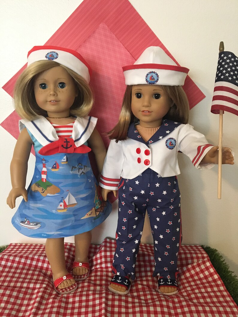 May include: Two dolls dressed in nautical-themed outfits. One doll wears a blue dress with a sailboat print, a red bow, and a white sailor hat. The other doll is in a white jacket, blue star-patterned pants, and a sailor hat. An American flag is held by one doll.
