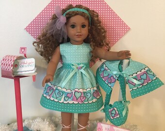 May include: A doll wearing a turquoise dress with white polka dots and a heart-themed skirt. Accessories include a matching bolero, purse, headband, and pink ballet flats. A decorative mailbox and cards with text complete the ensemble.
