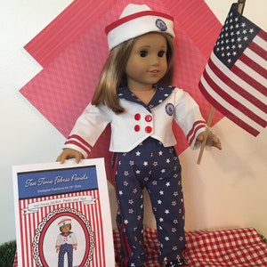 May include: A doll dressed in a sailor-themed outfit, including a white jacket with red buttons, navy pants with white stars, and a matching hat. The outfit is paired with anchor-decorated shoes. A small American flag is held nearby.