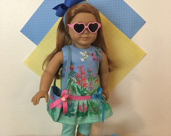 May include: A doll wearing a blue and green floral dress with a pink and blue bag, blue leggings, and pink heart-shaped sunglasses. The doll is standing on a green surface with a blue and green pillow with a floral design.