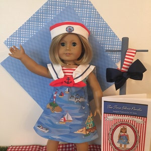 May include: A doll wearing a sailor-themed dress and hat. The blue dress features a nautical print with sailboats and islands. The white hat has a red band and a nautical emblem. A matching bow and a pattern sheet are also visible.