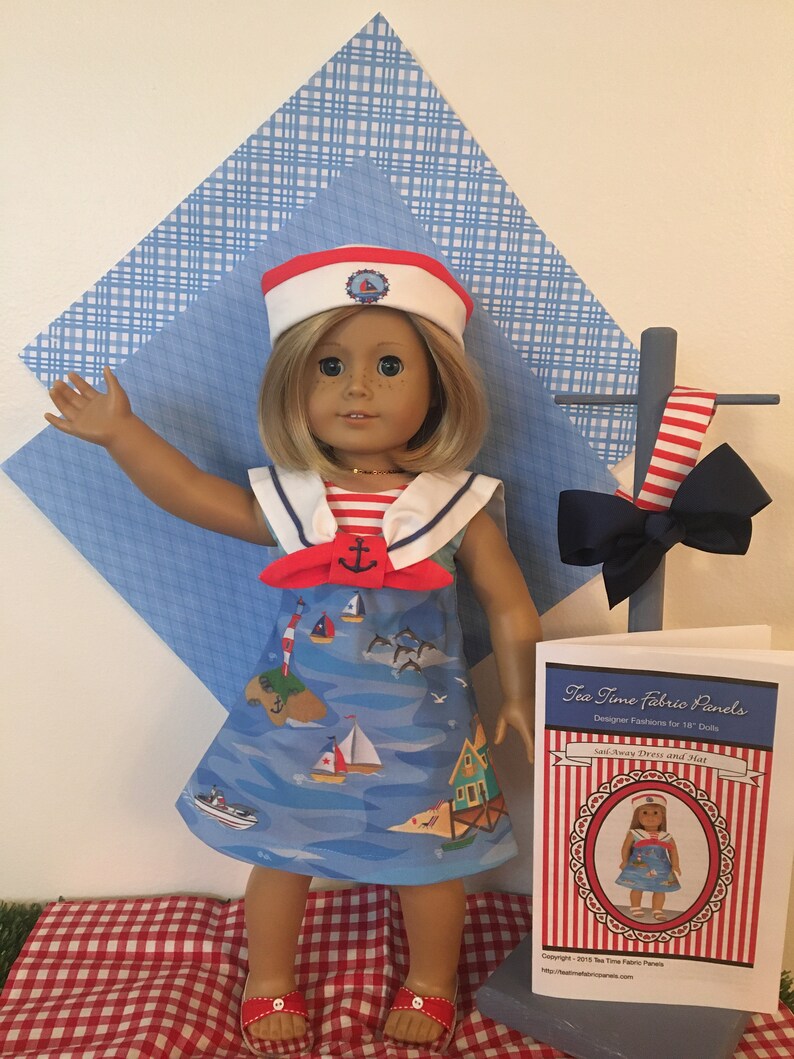 May include: A doll wearing a sailor-themed dress and hat. The blue dress features a nautical print with sailboats and islands. The white hat has a red band and a nautical emblem. A matching bow and a pattern sheet are also visible.