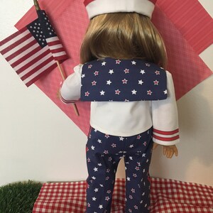 May include: A doll dressed in a patriotic sailor outfit, holding a small American flag. The outfit includes a white sailor hat with a red band, a white top with a navy blue collar adorned with white stars, and navy blue pants with white stars.