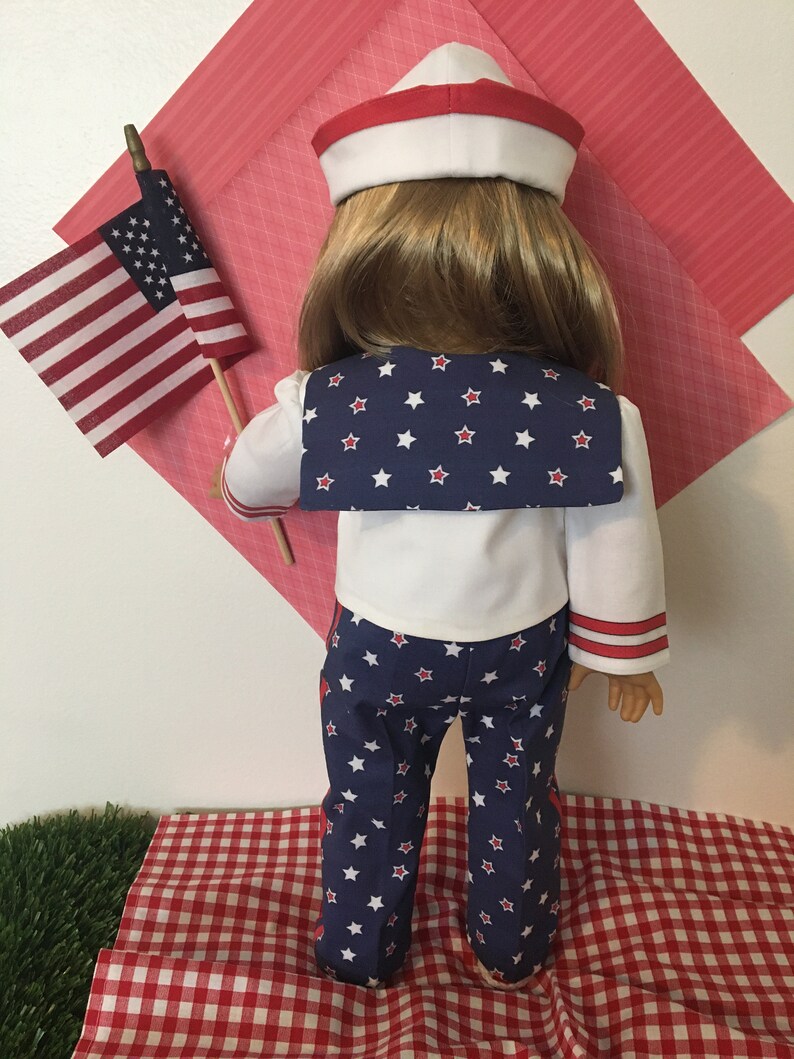 May include: A doll dressed in a patriotic sailor outfit, holding a small American flag. The outfit includes a white sailor hat with a red band, a white top with a navy blue collar adorned with white stars, and navy blue pants with white stars.