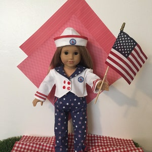 May include: A doll dressed in a sailor outfit, holding a small American flag. The outfit includes a white jacket with red accents and navy blue pants with white stars. The doll wears a white sailor hat with a red brim.