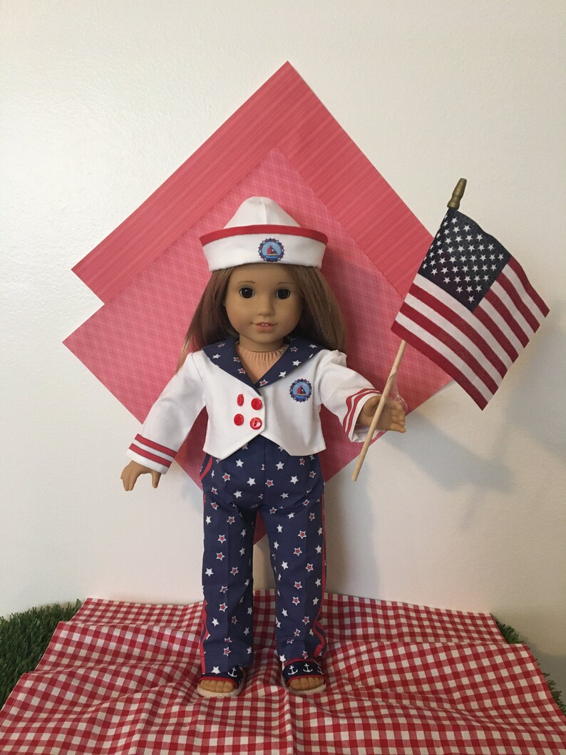 May include: A doll dressed in a sailor outfit, holding a small American flag. The outfit includes a white jacket with red accents and navy blue pants with white stars. The doll wears a white sailor hat with a red brim.