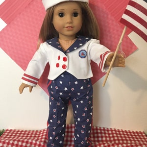 May include: A doll dressed in a sailor outfit with a white jacket, blue pants with white stars, and a matching hat. The outfit has red accents and a small emblem. The doll is holding a small American flag.