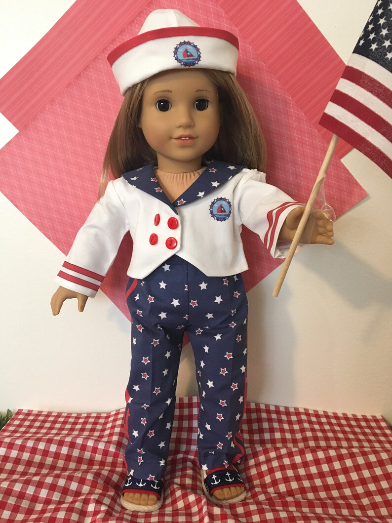 May include: A doll dressed in a sailor outfit with a white jacket, blue pants with white stars, and a matching hat. The outfit has red accents and a small emblem. The doll is holding a small American flag.