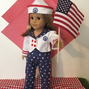 May include: A doll dressed in a sailor outfit, holding an American flag. The outfit includes a white jacket with red buttons, blue star-patterned pants, and a white sailor hat with a red trim. The doll is standing on a red and white checkered surface.