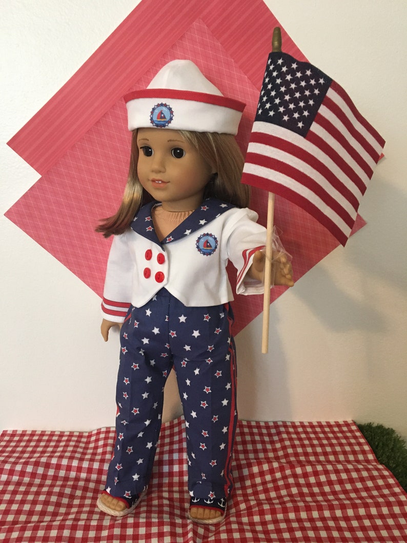 May include: A doll dressed in a sailor outfit, holding an American flag. The outfit includes a white jacket with red buttons, blue star-patterned pants, and a white sailor hat with a red trim. The doll is standing on a red and white checkered surface.