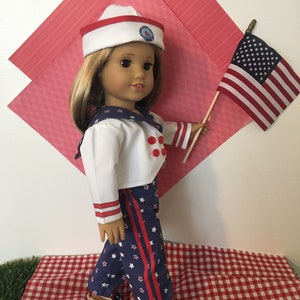 May include: A doll dressed in a sailor outfit, holding a small American flag. The outfit includes a white jacket with red buttons, blue pants with white stars and red stripes, and a sailor hat. The doll is standing on a red and white checkered surface.