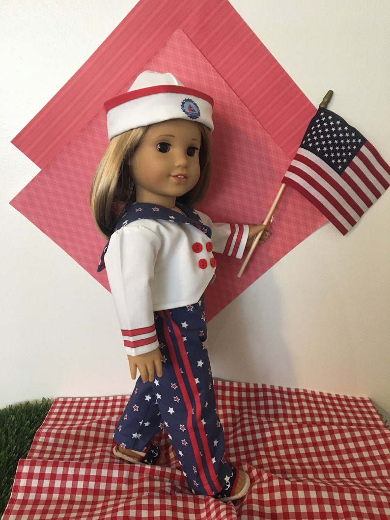 May include: A doll dressed in a sailor outfit, holding a small American flag. The outfit includes a white jacket with red buttons, blue pants with white stars and red stripes, and a sailor hat. The doll is standing on a red and white checkered surface.
