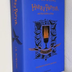 May include: A blue hardback book titled Harry Potter and the Goblet of Fire by J.K. Rowling. The cover features orange text, a central illustration of a flaming goblet, and the word Ravenclaw. The spine also displays the title and author.
