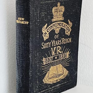 Puede incluir: Un libro del Nuevo Testamento de tapa dura negra con texto e imágenes en relieve dorados en la portada. El texto dice: IN COMMEMORATION OF SIXTY YEARS REIGN V.R. 1837-1897, acompañado de un icono de corona y una ilustración de un libro abierto.