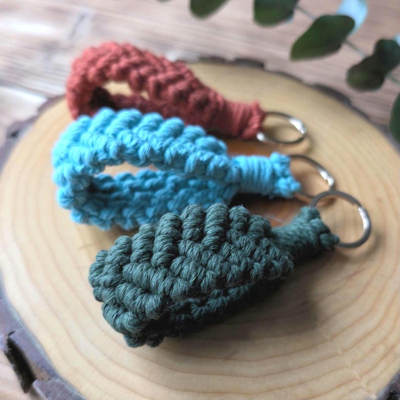 May include: Three macrame keychains in shades of rust, turquoise, and forest green. Each keychain features a woven design and a silver-coloured metal ring. The keychains are displayed on a light brown wooden surface.
