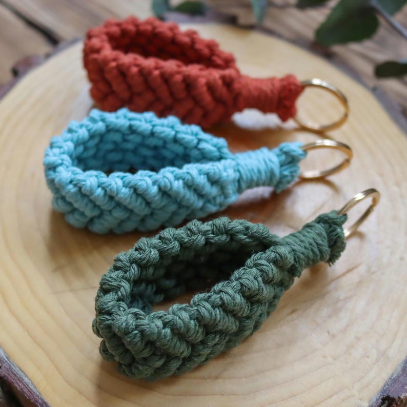 May include: Three macrame keyrings in shades of rust, teal, and olive green. Each keyring features a woven loop design with a gold-coloured metal ring. The keyrings are displayed on a light brown wooden surface.