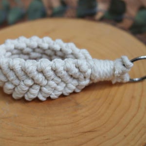 May include: An off-white macrame keychain with a woven pattern and a silver-coloured metal ring. The item is displayed on a wooden surface, highlighting the texture of the macrame. The keychain is a handcrafted item.