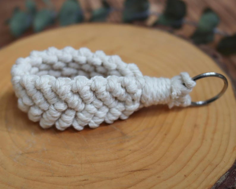 May include: An off-white macrame keychain with a woven pattern and a silver-coloured metal ring. The item is displayed on a wooden surface, highlighting the texture of the macrame. The keychain is a handcrafted item.