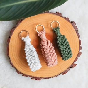 Puede incluir: Tres llaveros de macramé en blanco, rosa y verde, cada uno con un anillo dorado. Los llaveros están dispuestos sobre una rodaja de madera, con una hoja verde en la esquina superior izquierda. Los llaveros tienen un diseño tejido y texturizado.