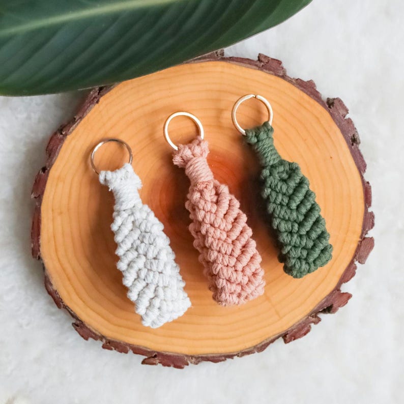 May include: Three macrame keyrings in white, pink, and green, each with a gold-coloured ring. The keyrings are arranged on a wooden slice, with a green leaf in the upper left corner. The keyrings have a woven, textured design.