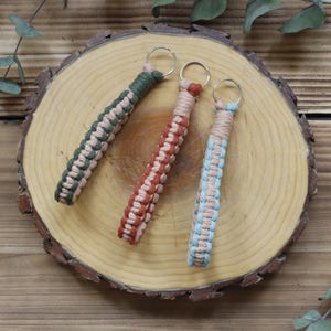 Pu&ograve; includere: Tre portachiavi in macram&egrave; verde, ruggine e blu con accenti beige, ciascuno attaccato a un portachiavi argentato. I portachiavi sono esposti su una fetta di legno, con uno sfondo di venature naturali del legno e rametti di verde.