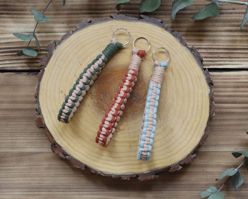 Pu&ograve; includere: Tre portachiavi in macram&egrave; verde, ruggine e blu con accenti beige, ciascuno attaccato a un portachiavi argentato. I portachiavi sono esposti su una fetta di legno, con uno sfondo di venature naturali del legno e rametti di verde.