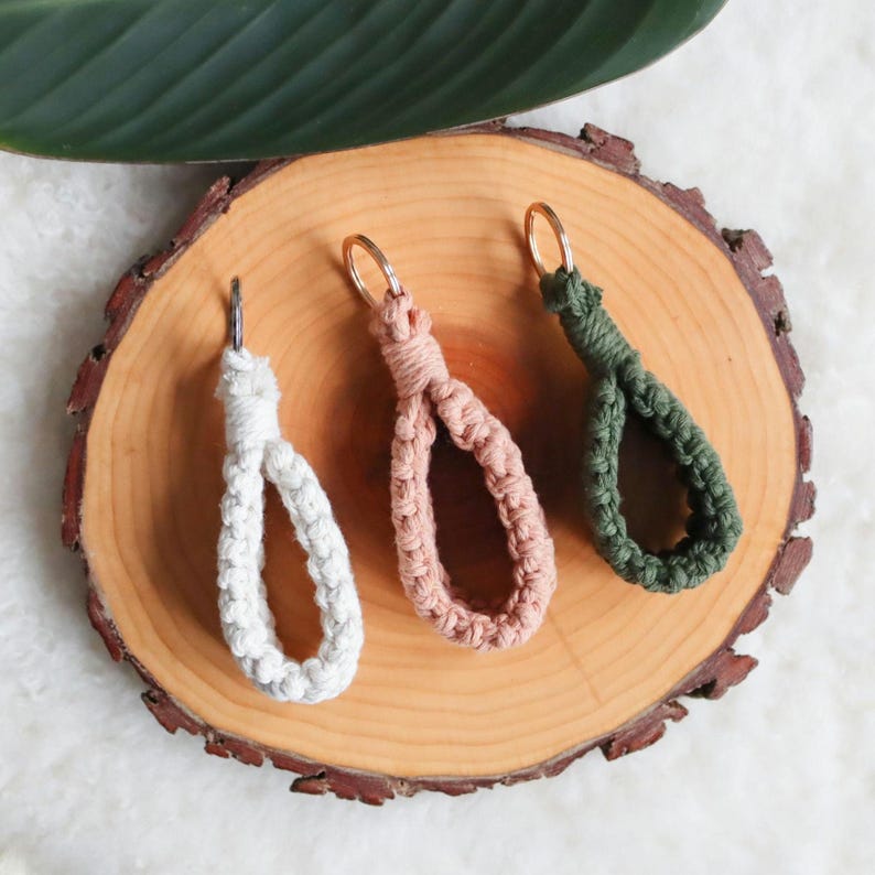 May include: Three macrame keyrings in white, blush pink, and olive green, each with a gold-coloured ring. The keyrings are displayed on a round wooden slice with a natural bark edge, with a green leaf in the background.