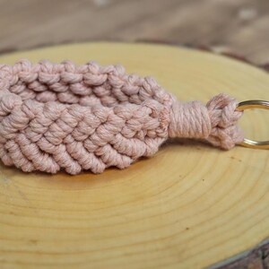 May include: A handmade macram&eacute; keyring in a soft, dusty rose colour. The woven design features a textured pattern, with a gold-coloured metal keyring attached. The keyring is displayed on a wooden surface.