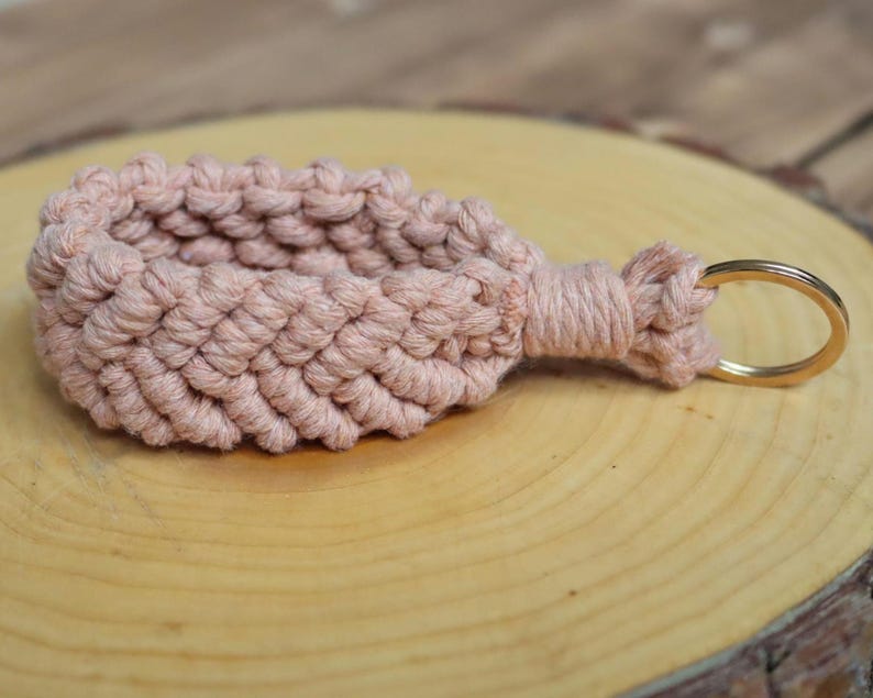 May include: A handmade macram&eacute; keyring in a soft, dusty rose colour. The woven design features a textured pattern, with a gold-coloured metal keyring attached. The keyring is displayed on a wooden surface.