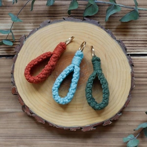May include: Three macrame keychains in shades of red-orange, light blue, and green, each with a gold-coloured metal ring. The keychains are arranged on a wooden slice with a natural edge, showcasing a handmade craft.