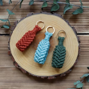 May include: Three macrame keyrings in shades of red, blue, and green, each with a gold-coloured ring. The keyrings are arranged on a wooden slice, with a natural wood grain background and sprigs of greenery.