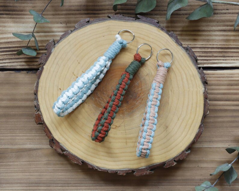 Pu&ograve; includere: Tre portachiavi in macram&egrave; nei toni del blu, bianco, verde, arancione e beige. I portachiavi sono disposti su una fetta di legno, con un motivo a venature naturali. Ogni portachiavi ha un anello di metallo.