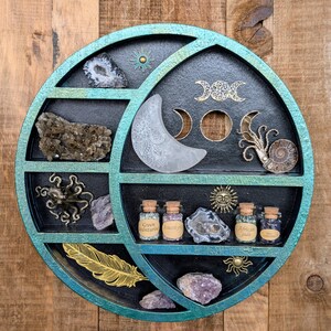 May include: A round, multi-compartment shelf with a teal and black finish, displaying crystals, decorative elements, and small glass bottles. The shelf features a crescent moon, sun, and other celestial designs. The bottles are labeled with the names of the crystals.