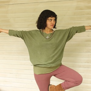 May include: A woman wearing a green, long-sleeved shirt and pink pants stands on one leg with her other leg bent at the knee. She is wearing brown boots and a silver necklace.