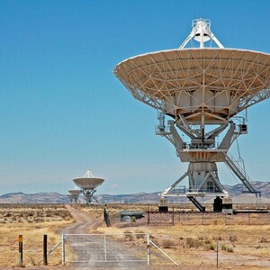 May include: A large white satellite dish antenna in a desert landscape. The antenna is pointed towards the sky and is surrounded by a fence. There are two smaller antennas in the distance.