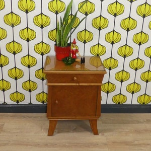 Könnte beinhalten: Ein Nachttisch aus Holz mit einer Glasplatte und einer Schublade. Der Tisch ist hellbraun und hat ein schlichtes Design. Er steht auf einem Holzboden vor einer Wand mit einem gelben und schwarzen geometrischen Muster.