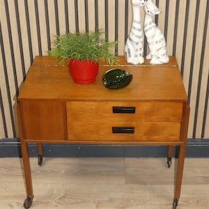 May include: A mid-century wooden side table with two drawers and a small cabinet. The table is finished in a reddish-brown hue and has four legs with castors. A red plant pot, a green glass ashtray, and two ceramic cat ornaments are on top.