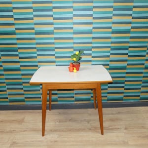 Könnte beinhalten: Ein Retro-Beistelltisch mit weißer Tischplatte und Holzbeinen. Eine kleine Topfpflanze mit gelben Früchten und eine rote Figur stehen auf dem Tisch. Der Hintergrund zeigt eine farbenfrohe gestreifte Tapete.