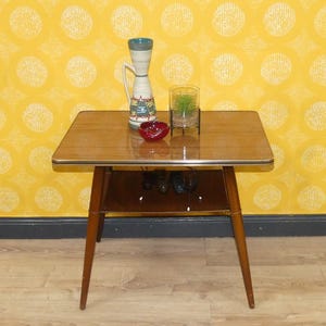May include: A vintage wooden side table with a rectangular top and a lower shelf. The table has a light brown finish with a gold trim. A decorative vase, a small red bowl, and a glass with a plant are on the table.